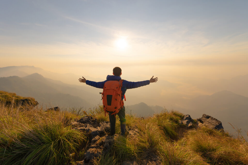 A person with an orange backpack stands on a grassy mountain peak at sunrise, embracing the misty Canadian landscape with arms outstretched capturing the sense of renewal, freedom, and the importance of sleep health.