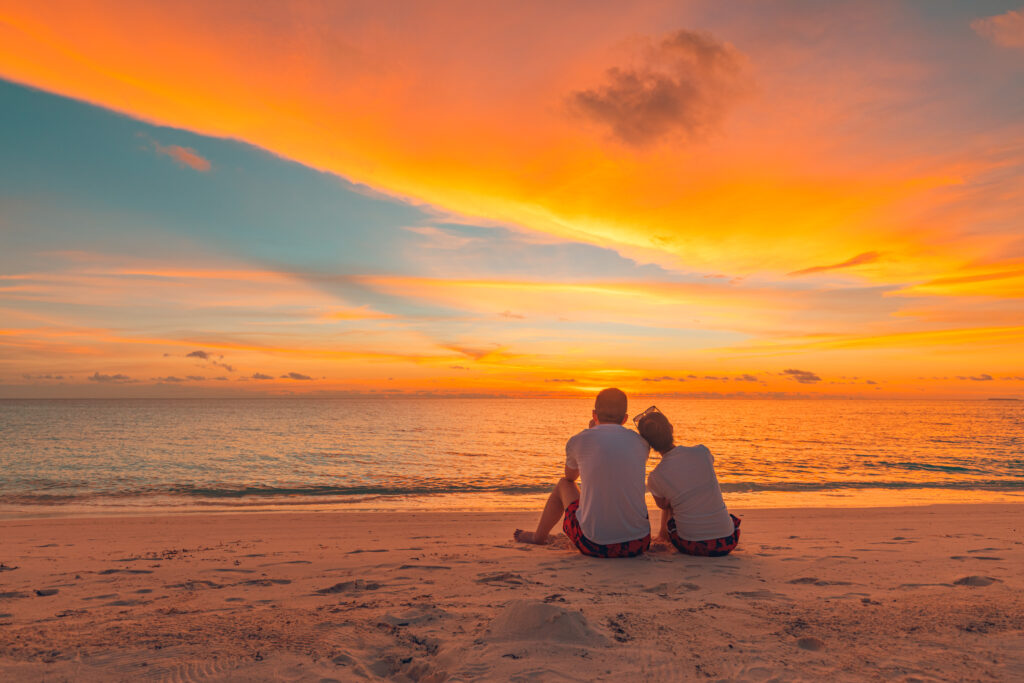 Deux personnes sont assises l'une près de l'autre sur une plage de sable, face à l'océan et à un ciel orange et jaune vibrant, créant une atmosphère paisible et romantique.