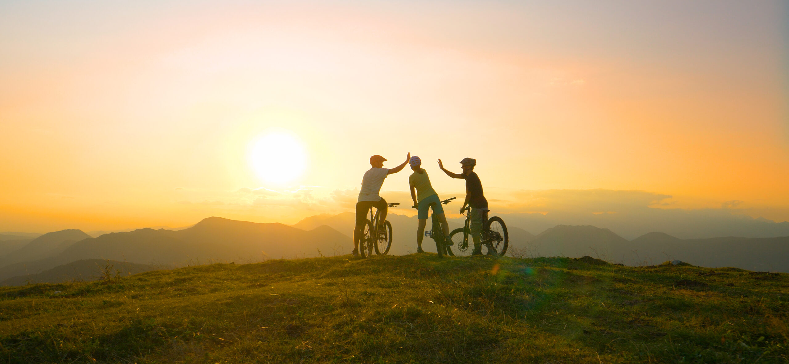 Three people with bicycles giving high-fives on a grassy hilltop at sunset, celebrating overcoming a sleep disorder, with mountains and a bright sun in the background.