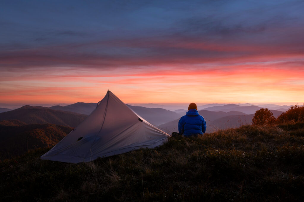 Une personne portant une veste bleue est assise à côté d'une tente sur une colline herbeuse, recherchant le calme au milieu des chaînes de montagnes au coucher du soleil - un cadre paisible qui pourrait soulager l'insomnie ou offrir un soulagement au stress d'un trouble du sommeil.