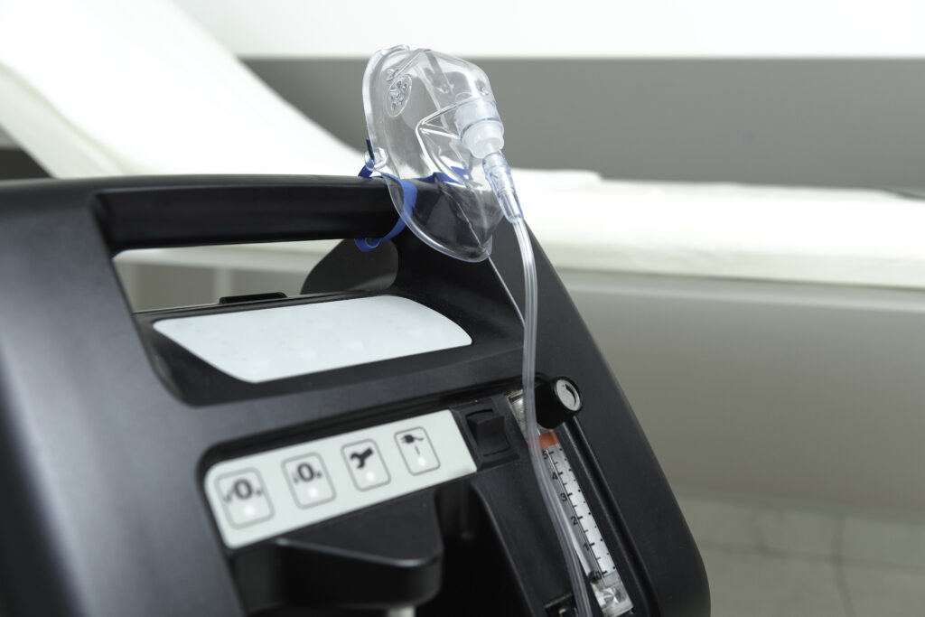A close-up of an oxygen concentrator machine with an attached transparent oxygen mask, used for oxygen therapy, positioned in a clinical or hospital setting with a white patient bed in the background.