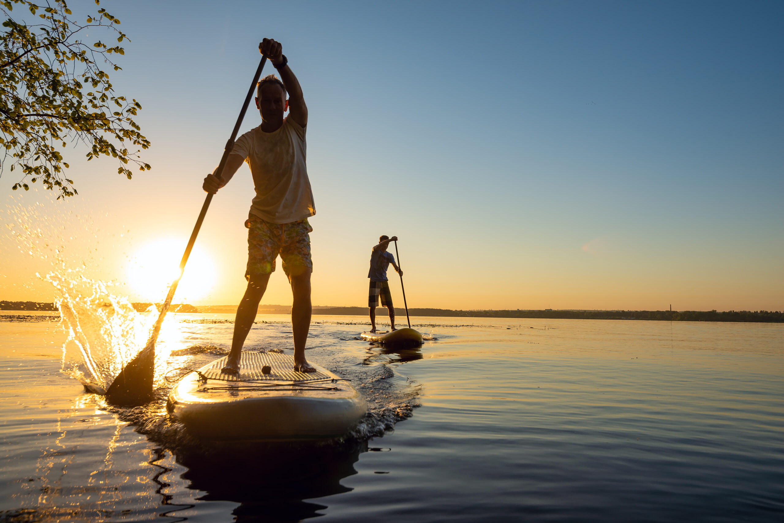 Deux personnes font du paddleboard sur un plan d'eau calme au coucher du soleil, avec une personne au premier plan qui s'amuse à éclabousser l'eau - une scène idyllique qui souligne l'importance de la relaxation et du soulagement du stress dans la gestion d'un trouble du sommeil tel que l'apnée du sommeil.