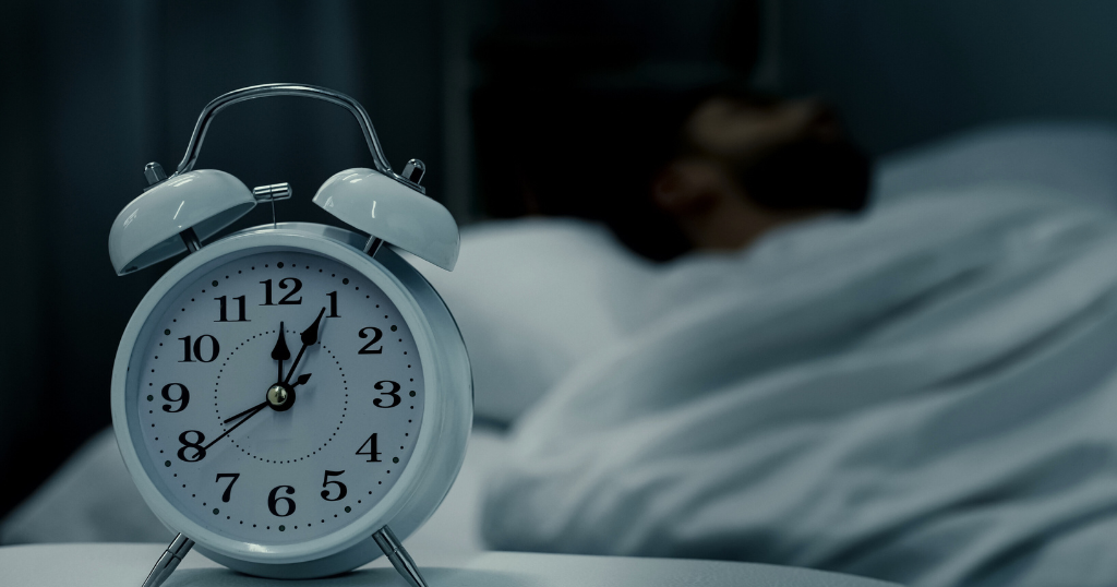 Alarm clock on bedside table with person sleeping, promoting consistent sleep schedule.