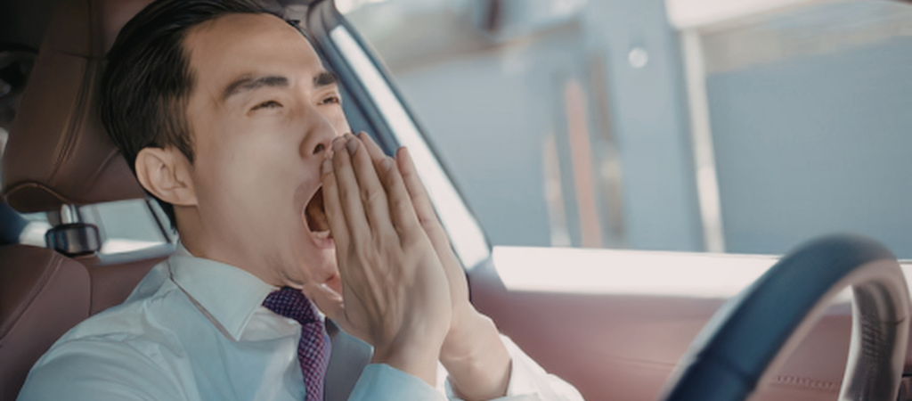 Une personne qui bâille alors qu'elle est assise au volant d'une voiture, illustrant la somnolence diurne et la fatigue liées à des troubles du sommeil ou à des causes hormonales.