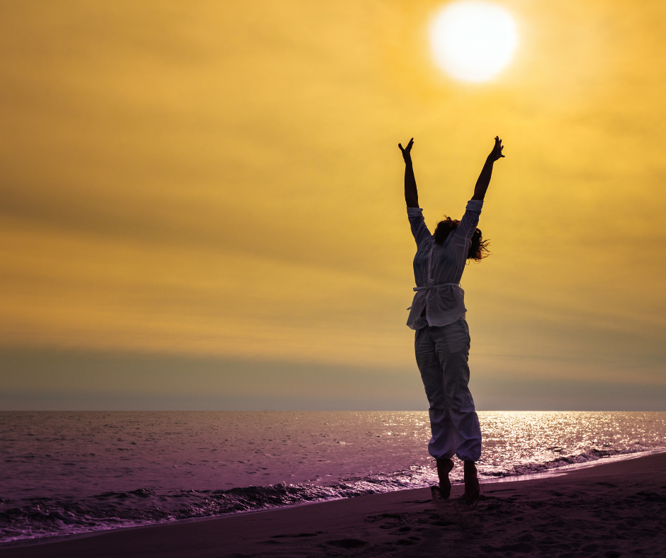 Une personne debout sur une plage au coucher du soleil, les bras levés, symbolisant un meilleur bien-être mental grâce à un sommeil réparateur.