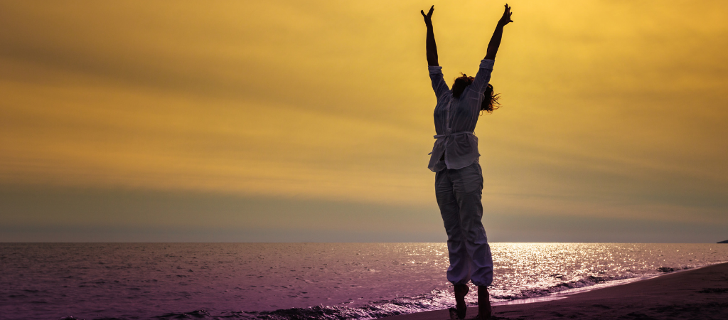 Person standing on a beach at sunset with arms raised, representing improved mental well‑being and balance through healthy sleep.