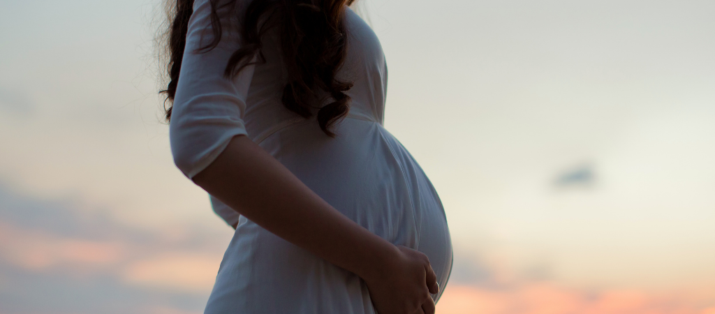 Pregnant person standing outdoors at sunset with a hand resting on the abdomen, representing pregnancy‑related fatigue and sleep changes.