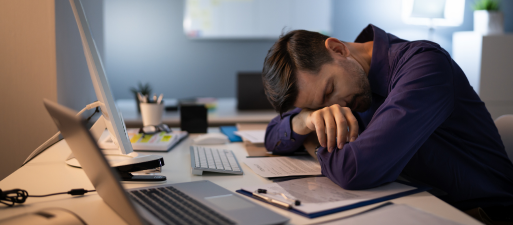 Un employé de bureau endormi à son bureau, entouré d'un ordinateur et de documents, illustrant la fatigue diurne et la somnolence excessive, symptômes courants de l'apnée du sommeil souvent négligés.